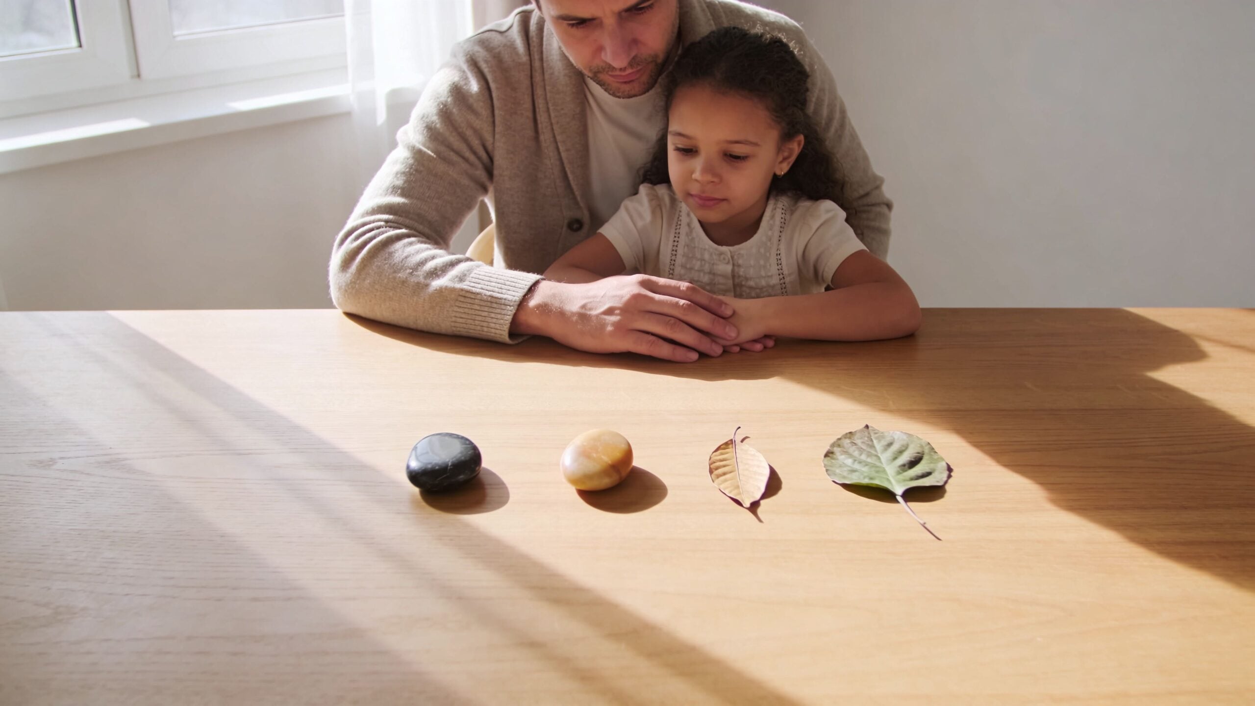 A father and his young daughter sitting together at a wooden table looking at natural objects.