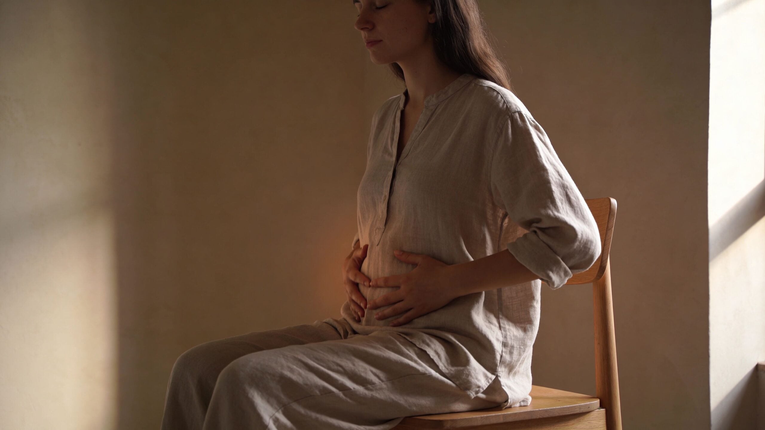 A pregnant woman sitting on a wooden chair, holding her belly with both hands in a calm manner.