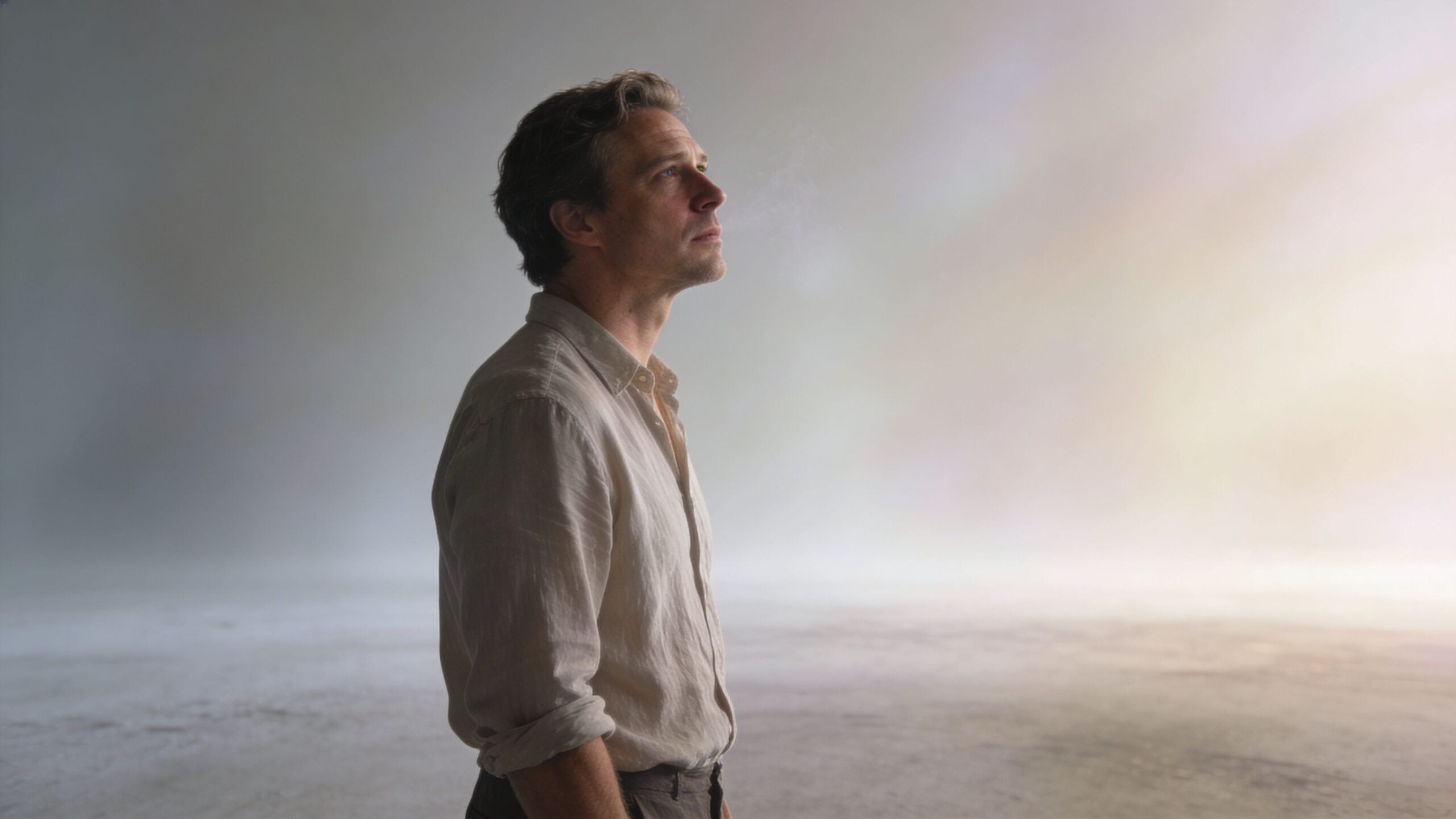 A contemplative man in a light shirt standing alone in a vast, hazy, and minimalist space.